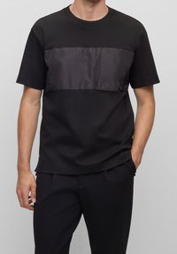 Muž v černém tričku s krátkým rukávem a lesklým látkovým panelem na hrudi a černých kalhotách, s rukama v kapsách na šedém pozadí.