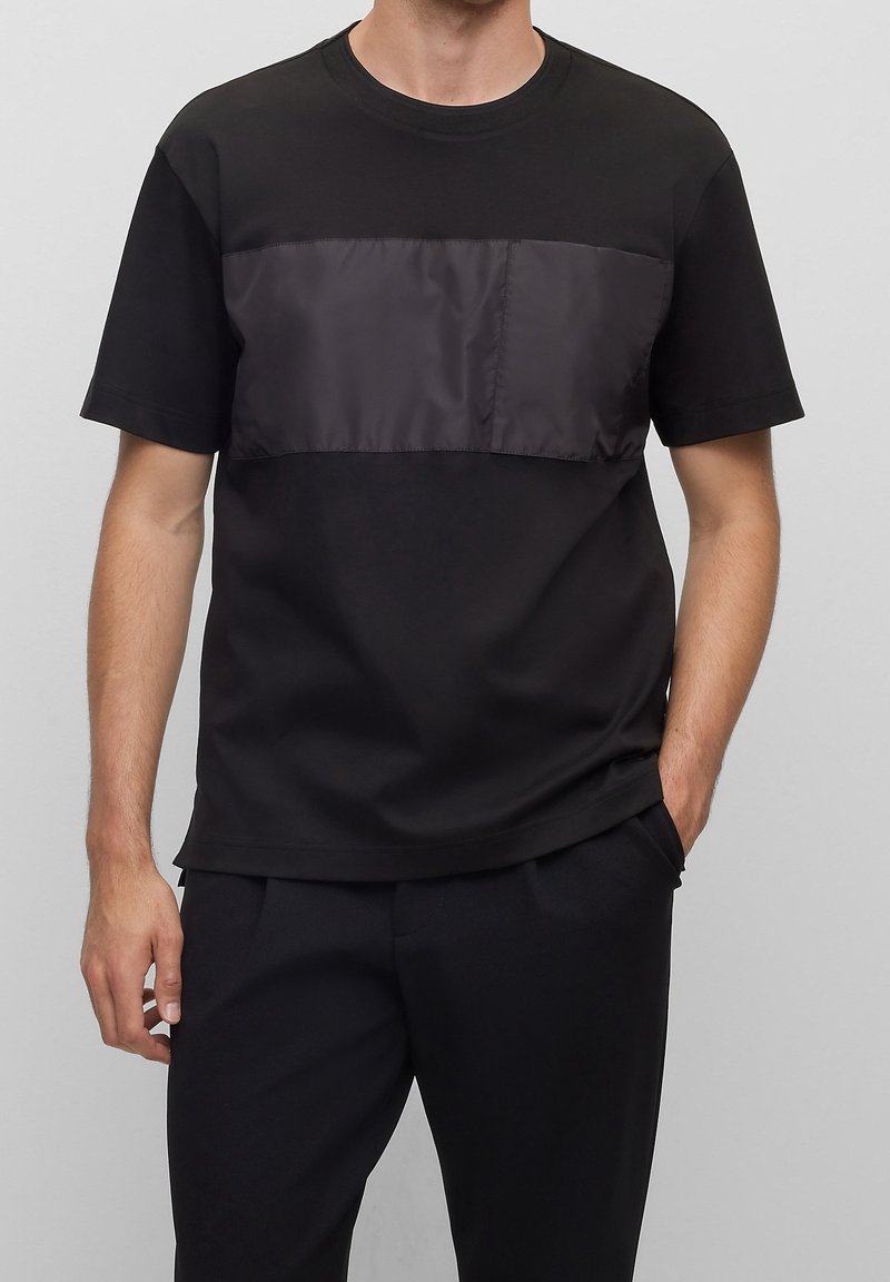 Muž v černém tričku s krátkým rukávem a lesklým látkovým panelem na hrudi a černých kalhotách, s rukama v kapsách na šedém pozadí.