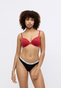 Červená podprsenka s výplňou a tenkými ramienkami, hladký povrch. Čierne tangá s bielym pásom s logom Calvin Klein. Jednoduchý, priliehavý dizajn.