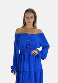 Blå off-shoulder kjole med lange ærmer, knappedetalje, smocket talje og lagdelt nederdel, lavet af letvægtsstof med en glat tekstur.