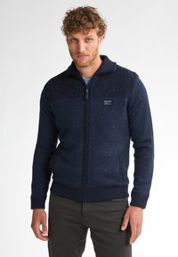 Marineblauwe gebreide trui met een gestructureerd bovenstuk en een gladde romp, voorzien van een ritssluiting en zijzakken. Neutrale spikkels doorheen.