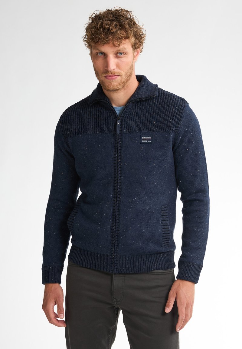Marineblauwe gebreide trui met een gestructureerd bovenstuk en een gladde romp, voorzien van een ritssluiting en zijzakken. Neutrale spikkels doorheen.
