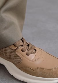 Sneaker marrone caratterizzato da un mix di pelle testurizzata e rete, punta rotonda, lacci piatti e suola bianca imbottita; abbinato a pantaloni khaki.