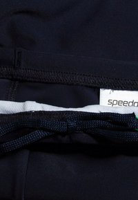 Pantaloni da bagno neri con fodera interna bianca, dotati di chiusura con cordino di colore blu scuro. Etichetta del marchio Speedo visibile.