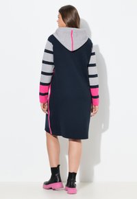 Abito lavorato a maglia blu navy con maniche lunghe a strisce grigie e rosa, cappuccio con accento rosa e dettagli a toppa sui lati; abbinato a stivali platform neri e rosa.