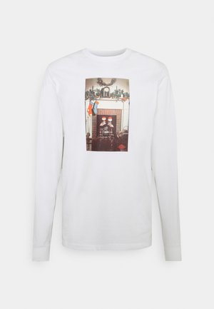 T-shirt blanc à manches longues avec un imprimé central représentant une scène festive de cheminée avec des bas de Noël et des éléments décoratifs.