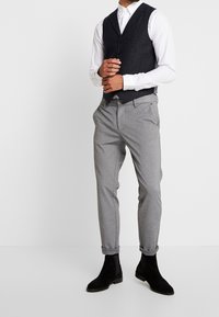 Pantalones gris ajustados combinados con un chaleco de tartán oscuro sobre una camisa blanca, complementados con botas de ante negras hasta el tobillo.