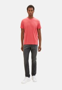 Korallenfarbenes T-Shirt mit kurzen Ärmeln, kombiniert mit dunkelgrauen Jeans und weißen Sneakers. Einfaches Design mit Rundhalsausschnitt. Stoffgemisch aus Baumwolle.