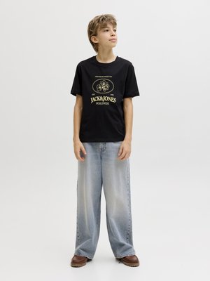 Chłopiec o krótkich jasnobrązowych włosach stoi, ubrany w czarną koszulkę z nadrukiem, luźne jasnoniebieskie dżinsy i brązowe buty, na tle jednolitego jasnego tła.