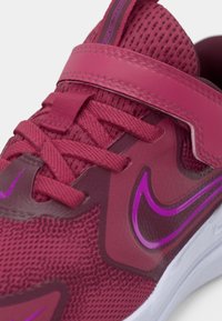 Scarpa da ginnastica rossa con una tomaia in rete testurizzata, finiture sintetiche lisce e dettagli viola sul logo e sulla cinghia. Presenta lacci e una cinghia in velcro.