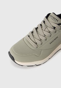 Nærbilde av beige Skechers-sneaker med perforert tå, tykke lisser, svart og hvit såle, og Skechers-logo på siden og tungen.