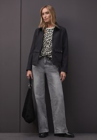 Schwarze Jacke mit zwei Fronttaschen über einem Oberteil mit Leopardenmuster, kombiniert mit grauen Jeans mit weitem Bein und schwarzen Stiefeletten. Schwarze Handtasche in der Hand.