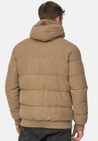 Chaqueta tipo puff en color marrón con costuras horizontales, capucha y puños acanalados, con un acabado texturizado y un parche con el logo en la parte posterior.
