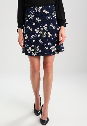 Mujer de pie con falda azul marino estampada con flores, camiseta negra de manga larga y zapatos de tacón altos negros con punta afilada sobre fondo blanco.