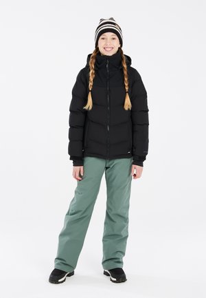 Schwarze, gepolsterte Jacke mit Reißverschluss und Kapuze; kombiniert mit lockeren grünen Hosen und schwarzen Winterstiefeln. Gestreifte Strickmütze unterstreicht den Stil.