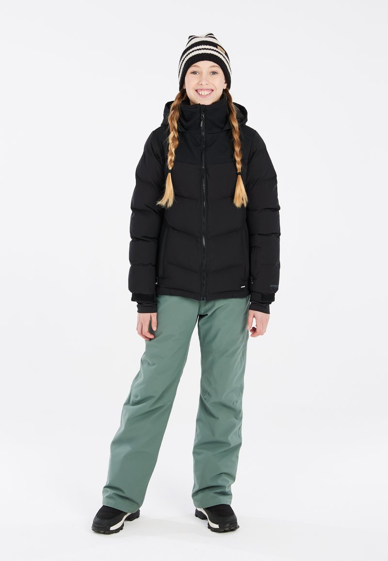 Doudoune noire, avec une fermeture éclair à l'avant et une capuche ; associée à un pantalon vert ample et des bottes d'hiver noires. Un bonnet tricoté rayé accentue le style.