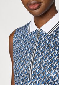 Zils polo kreklu ar baltu apkakli, aprīkots ar rāvējslēdzēju un ģeometrisku rakstu ar pelēkiem un gaiši zilajiem akcentiem.