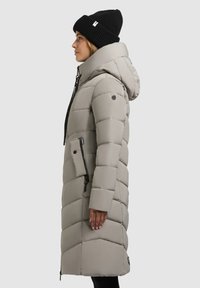 Abrigo de plumas gris claro con capucha, que presenta un bolsillo con cremallera, costuras diagonales y acentos negros. Combinado con un gorro de punto negro.