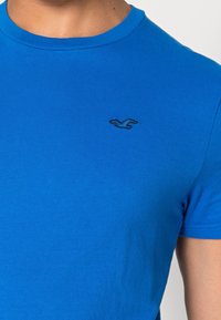 Modrý bavlněný tričko, krátké rukávy, žebrovaný kulatý výstřih, ma vykreslené malé černé vyšívané logo na levé hrudi. Hladká textura.