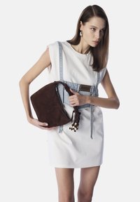 Bolso de ante marrón con forma curva y asa trenzada adornada con cuentas, sostenido por un modelo que lleva un vestido blanco y detalles de denim.