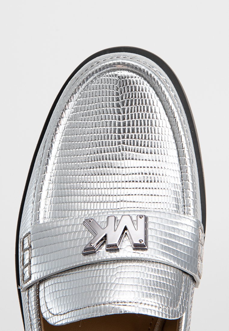 Mocassins métalliques argentés avec une finition texturée, arborant un accent de marque. La semelle est noire avec des bords lisses.