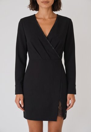 Donna che indossa un vestito nero a maniche lunghe modello avvolgente con scollatura a V e dettaglio in pizzo visibile sulla spacco frontale della coscia.