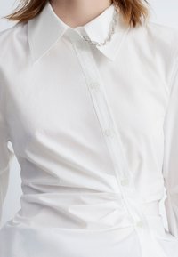 White button-up shirt with a classic collar, featuring textured fabric and a fitted waist. Includes clear buttons and subtle gathering details.