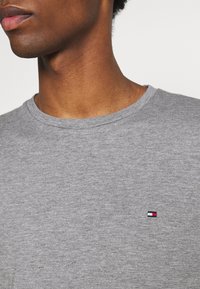 Šedé bavlněné tričko s kulatým výstřihem. Na hrudi je malé značkové logo v červené, bílé a modré barvě. Měkká, strukturovaná tkanina.