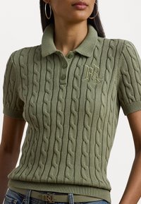 Polo-shirt en maille torsadée vert olive avec col côtelé, manches courtes et logo brodé. Texture douce et design ajusté avec un ourlet plat.