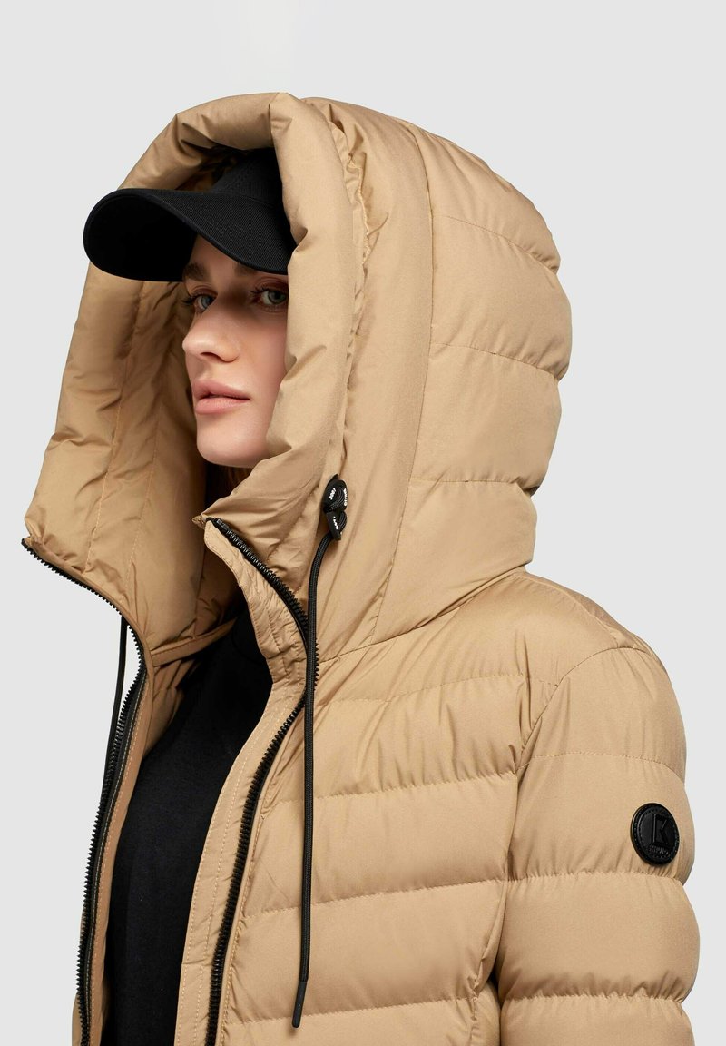 Beige Steppjacke mit großer Kapuze, gestepptem Design, schwarzem Reißverschluss und Kordeln. Kombiniert mit einer schwarzen Mütze.