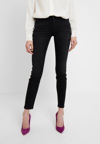 Mujer vistiendo jeans ajustados negros, blusa blanca de mangas largas y zapatos de tacón puntiagudos morados, de pie frente a un fondo blanco.