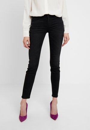 Mujer vistiendo jeans ajustados negros, blusa blanca de mangas largas y zapatos de tacón puntiagudos morados, de pie frente a un fondo blanco.