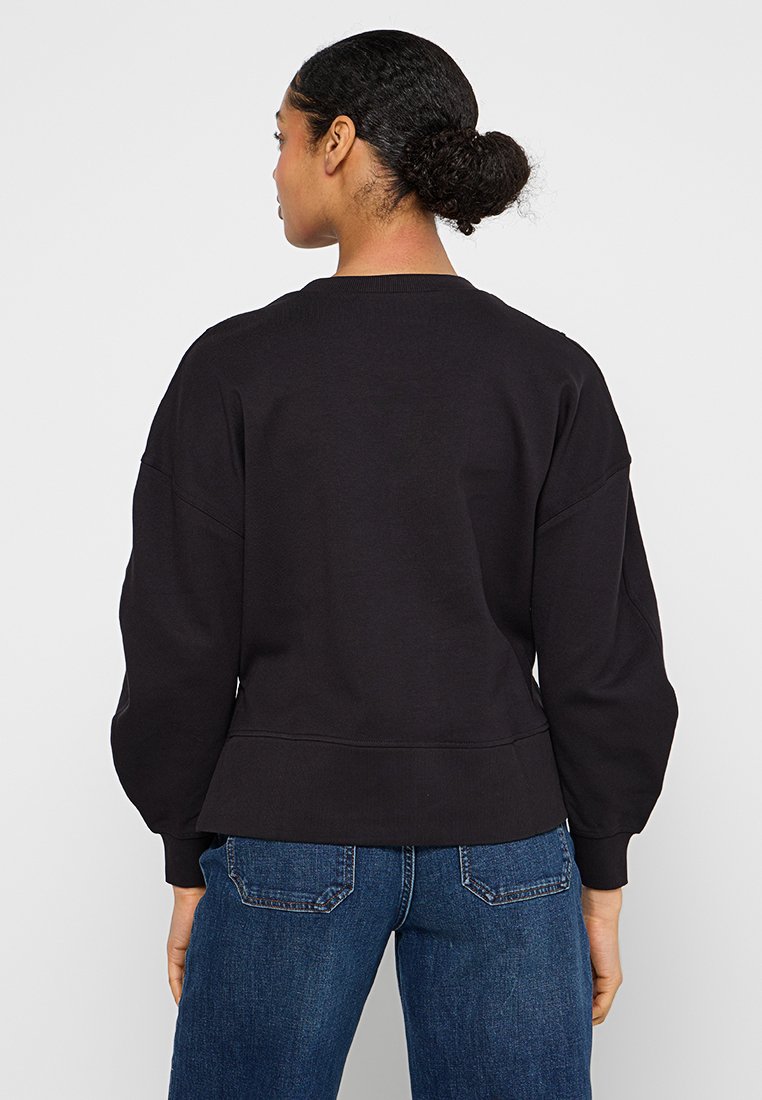 Czarny oversize sweter z okrągłym dekoltem i opuszczonymi ramionami. Materiał z francuskiej pętli i ściągacze u dołu. Noszony ze niebieskimi dżinsami.