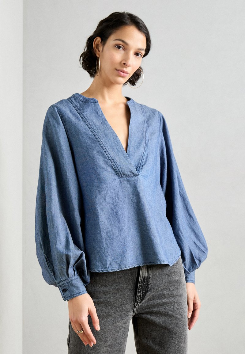 Denimbluse med dyp V-hals, lange puff-ermer og avslappet passform. Fargen er middels blå med en myk, teksturert overflate.