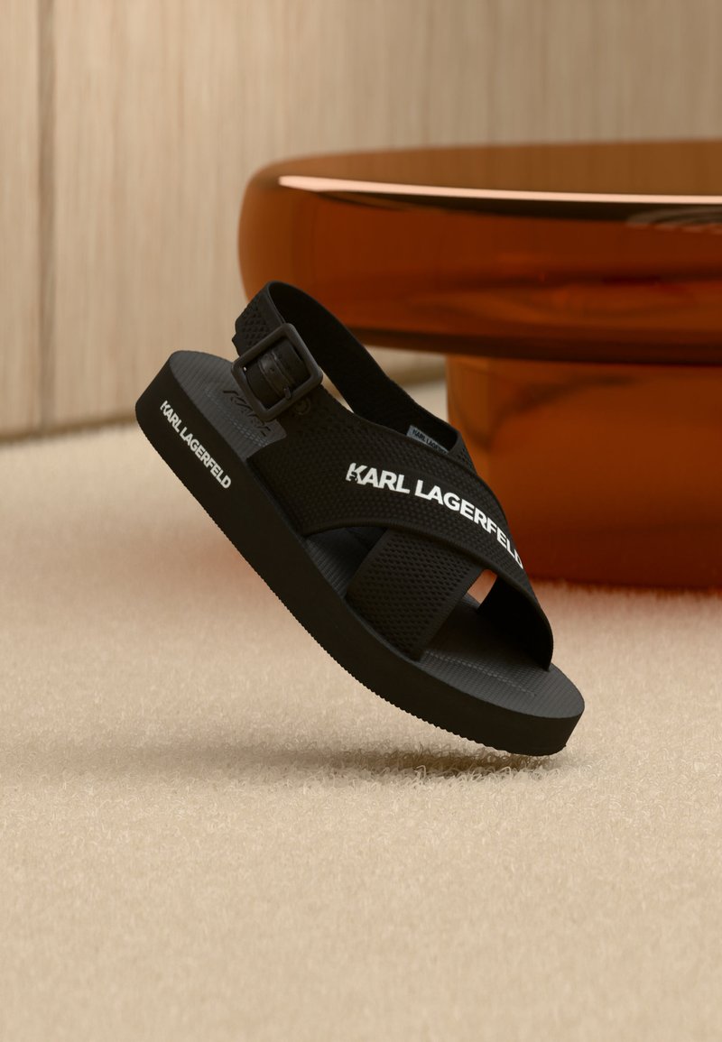 Μαύρο σανδάλι με ανοιχτή μύτη, ρυθμιζόμενο λουράκι, χοντρή σόλα και λογότυπο "KARL LAGERFELD" στις πλαϊνές λουρίδες και στην άκρη της σόλας.