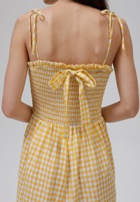Žluté kostkované šaty s nařaseným korzetem, volánkovým výstřihem a zavazovacími ramínky. Textura je lehká a po celém povrchu se nachází kostkovaný vzor.