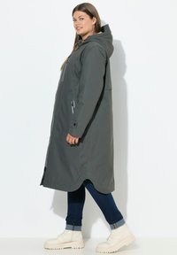 Abrigo largo gris impermeable con capucha, con bolsillos frontales, dobladillo curvado y cremalleras blancas. El modelo lleva puestos jeans azules y botas color crema.