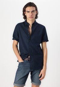 Chemise marine à manches courtes avec de petits pois blancs, fermeture boutonnée à l'avant, et coupe décontractée, associée à des shorts en denim avec ourlet roulé.