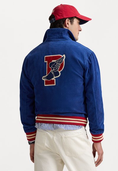 Blue corduroy jacket with a red and white striped ribbed hem, featuring a large embroidered winged ‘P’ logo on the back. Paired with a striped shirt.