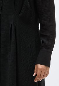 Μαύρο φόρεμα με υφή υφάσματος που έχει κάθετες ρυτιδώσεις. Μακριά μανίκια με φαρδιά μανσέτα. Λεπτομέρεια ενός χεριού που κρατά το ύφασμα.