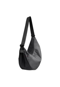 Sac bandoulière noir avec une large sangle réglable et une petite étiquette colorée sur le côté, présenté suspendu sur fond blanc.