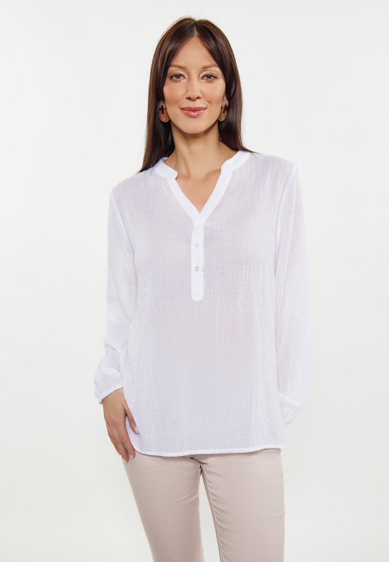 Weiße Bluse mit langen Ärmeln und strukturiertem Finish, mit knöpfbarem V-Ausschnitt und elastischen Bündchen, kombiniert mit hellen Hosen.