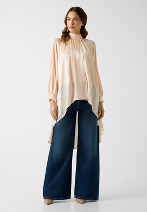 Frau trägt eine lockere, blassrosa Bluse mit hohem Kragen und langen Ärmeln sowie dunkle, weit geschnittene Jeans und steht vor einem schlichten Hintergrund.