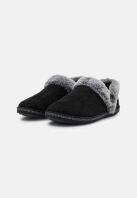 Svarta tofflor i mjuk mocka, med en grå syntetpälsfåll och en mönstrad sula med prickigt mönster. Rundad tådesign.