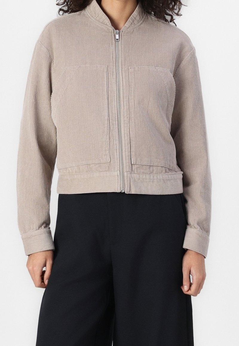 Beige corduroy jacket with a cropped design, front zip closure, and two chest pockets; features a round neckline and ribbed cuffs.