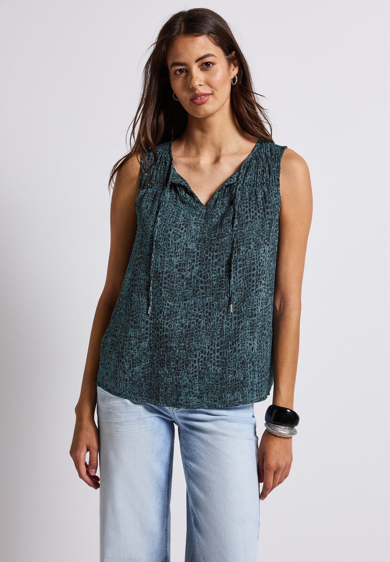 Sleeveless top in dark teal with a textured pattern, featuring a V-neck and drawstring detail. Paired with light blue wide-leg jeans.