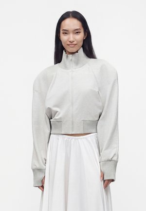Femme aux longs cheveux noirs portant un pull zippé gris clair aux épaules bouffantes et une jupe blanche fluide, debout devant un fond uni.