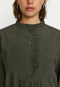 Blusa verde oliva con scollo arricciato, chiusura con bottoni e un tessuto texturizzato, caratterizzata da un design svasato e vita arricciata.