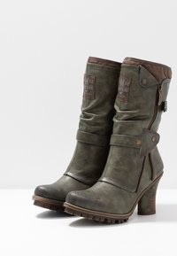 Botas verdes de tacón alto con cremallera y detalles de correa, que presentan un acento marrón en la parte superior y suelas gruesas, exhibidas sobre un fondo blanco.