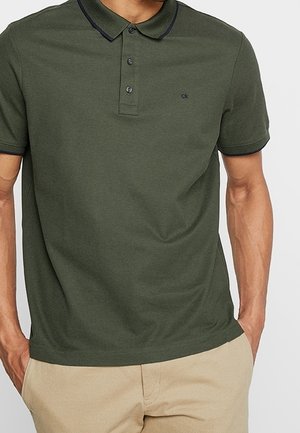 Mand iført olivengrøn kortærmet poloshirt med sort kant og beige bukser, hænder i lommerne, synlig fra skuldre til hofter.